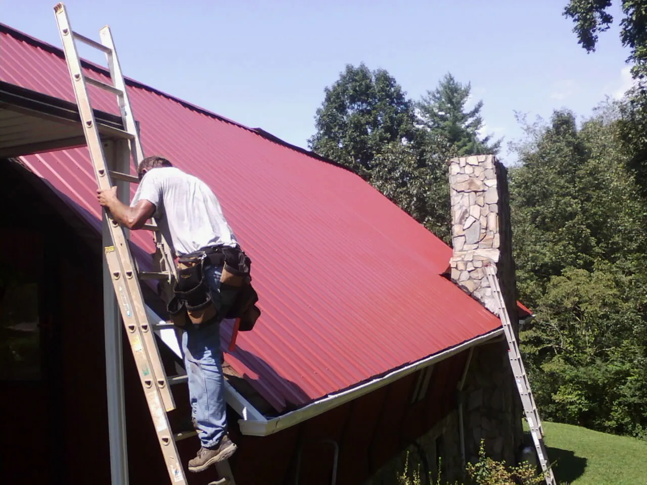 Roofer on a ladder beside a red metal roof and stone chimney — Soffit & Fascia Repair by Roofing Brothers in Annandale On Hudson